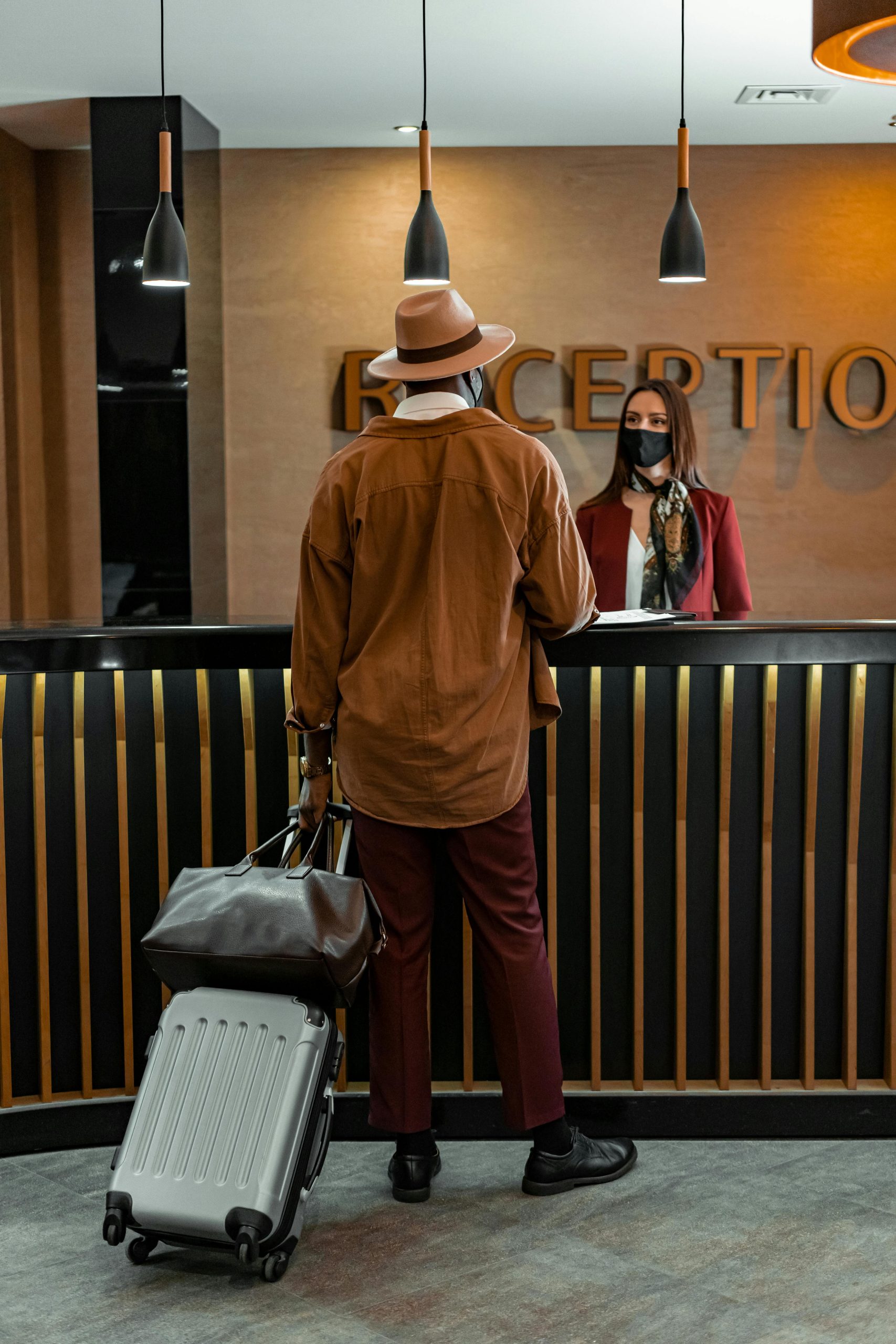 Man with luggage checking in at hotel reception. Staff wearing mask, indoors, travel theme.