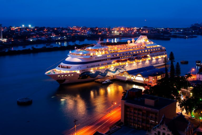 A brightly lit cruise ship docked at night, reflecting on calm river waters, surrounded by city lights.